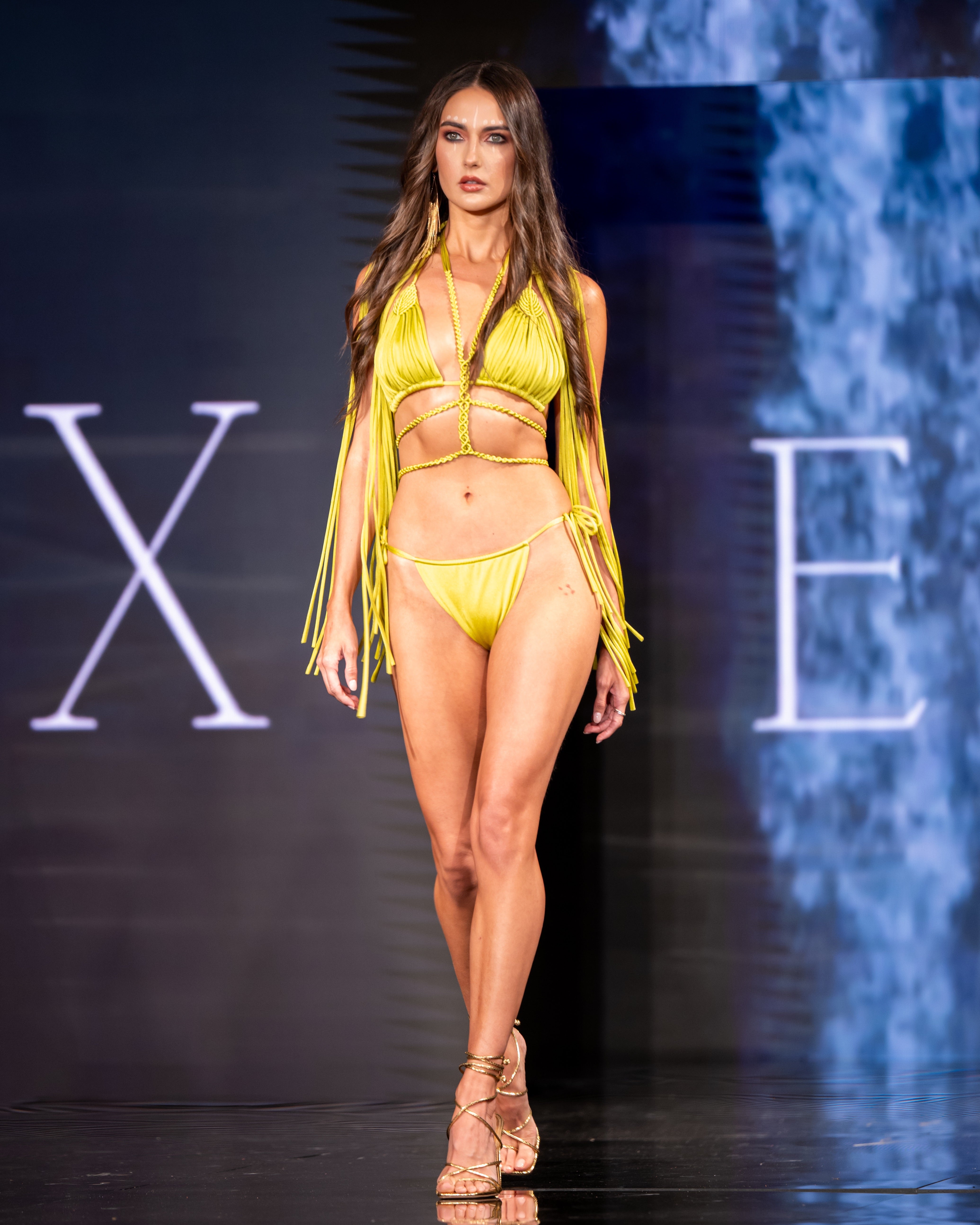 Model wearing a yellow fringe bikini set on a runway with a dark background.